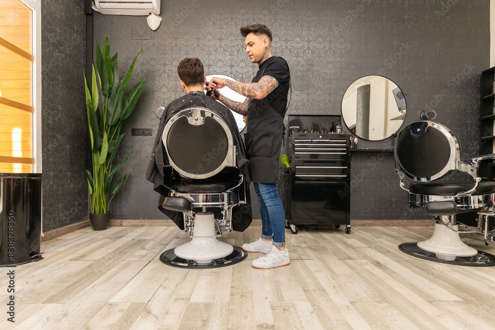 Fototapeta premium tattooed barber cutting the hair and beard of a young, dark-haired client.