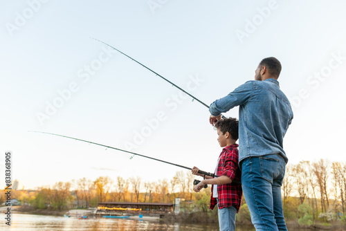 african american father and son fishing in the lake and sitting on wooden pier and holding fishing rod outdoors in summer, man teaches child to fish and relax outdoors on river