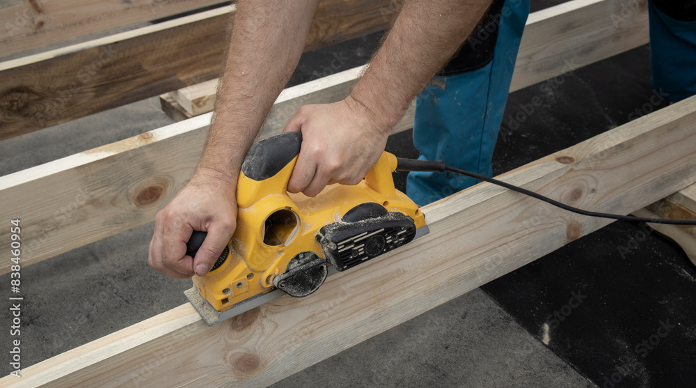 A close-up view of a carpenters hand using an electric planer to smooth a wooden beam. The tool is powered by a cord and the carpenter is carefully guiding it along the wood. A close-up view of a carpenters hand using an electric planer to smooth a wooden beam. The tool is powered by a cord and the carpenter is carefully guiding it along the wood.