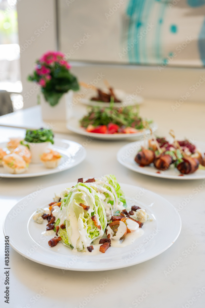 Wedge salad with bacon crumbles and white dressing on a white plate. Various other food on plates in background. Vertical color photo Wedge salad with bacon crumbles and white dressing on a white plate. Various other food on plates in background. Vertical color photo
