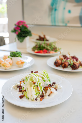 Wedge salad with bacon crumbles and white dressing on a white plate. Various other food on plates in background. Vertical color photo