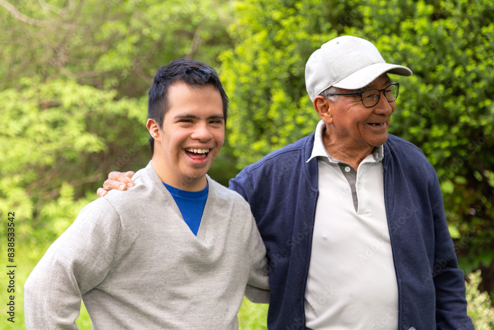 A man and a young man with Down syndrome are standing happily in a park. The young man is wearing a gray sweater. A man and a young man with Down syndrome are standing happily in a park. The young man is wearing a gray sweater.