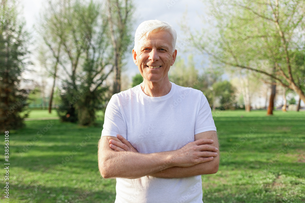 portrait of an old gray-haired male pensioner in a white T-shirt in the park, an elderly senior stands with his arms crossed and smiles outdoors portrait of an old gray-haired male pensioner in a white T-shirt in the park, an elderly senior stands with his arms crossed and smiles outdoors