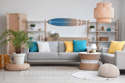 Light interior of living room with cozy sofa, coffee table, lamp and surfboard