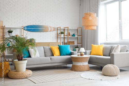 Light interior of living room with cozy sofa, coffee table, lamp and surfboard