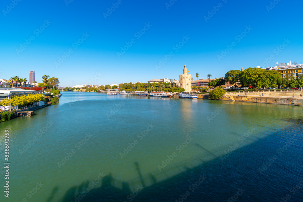 Obraz premium The Guadalquivier River as it runs through the historic center of Seville, Spain with the Triana district on one side, the Torre del Oro watchtower on the other, and the Sevilla Tower in the distance.
