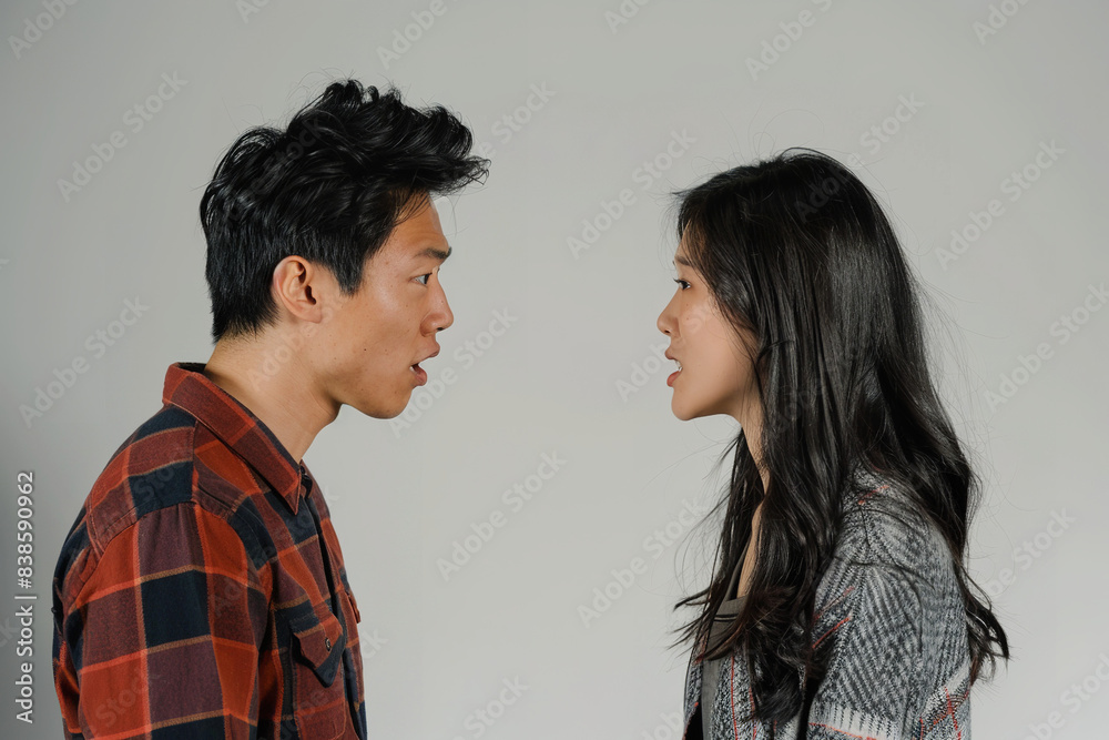 On a stark white background, an Asian young couple stands in tense confrontation, their expressions etched with anger and frustration On a stark white background, an Asian young couple stands in tense confrontation, their expressions etched with anger and frustration