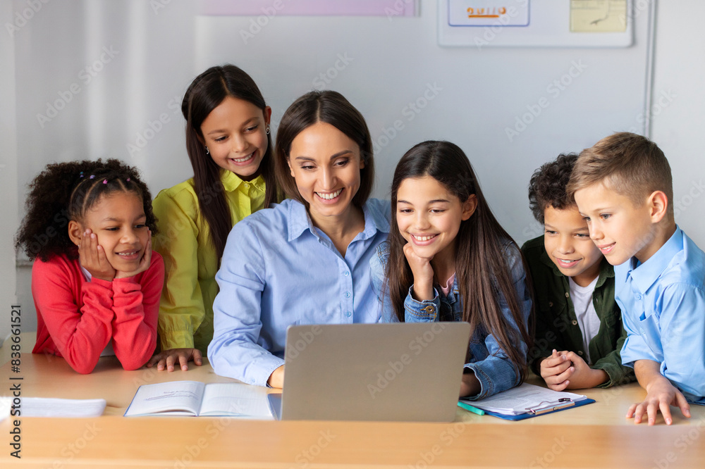 Happy young female tutor and excited diverse children smiling and looking at laptop screen while gathering around table during lesson at school Happy young female tutor and excited diverse children smiling and looking at laptop screen while gathering around table during lesson at school