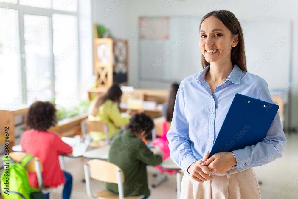 Portrait of cheerful European female teacher standing in classroom during lesson with pupils studying on background Portrait of cheerful European female teacher standing in classroom during lesson with pupils studying on background
