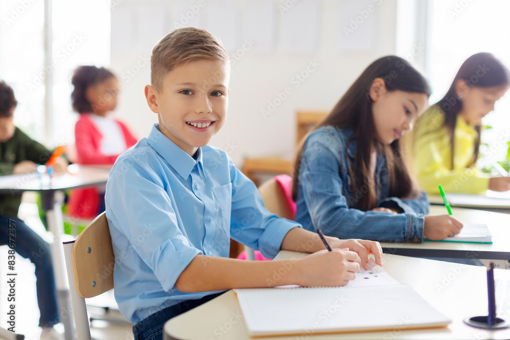 Portrait of positive European schoolboy sitting at his desk in classroom during lesson in secondary school and smiling at camera Portrait of positive European schoolboy sitting at his desk in classroom during lesson in secondary school and smiling at camera