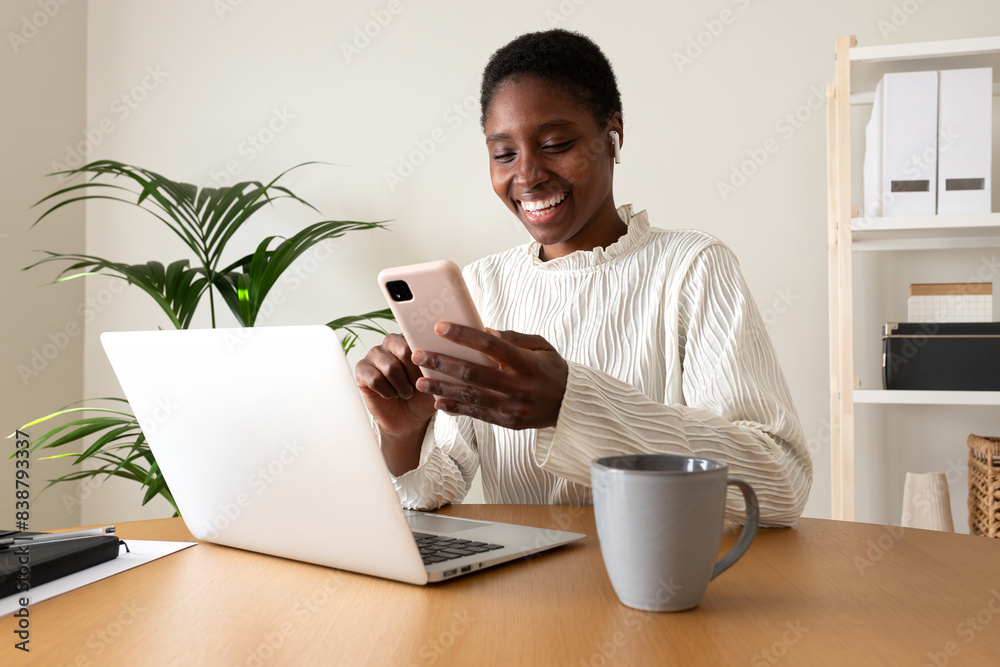 Young happy African American woman working at home office texting with mobile phone and using laptop. Young happy African American woman working at home office texting with mobile phone and using laptop.