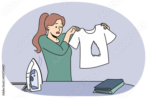Clumsy housewife ironing clothes burnt hole in t-shirt and dont know how to fix mistake. Woman stands near ironing board with clothes and iron and looks at damaged item in confusion.