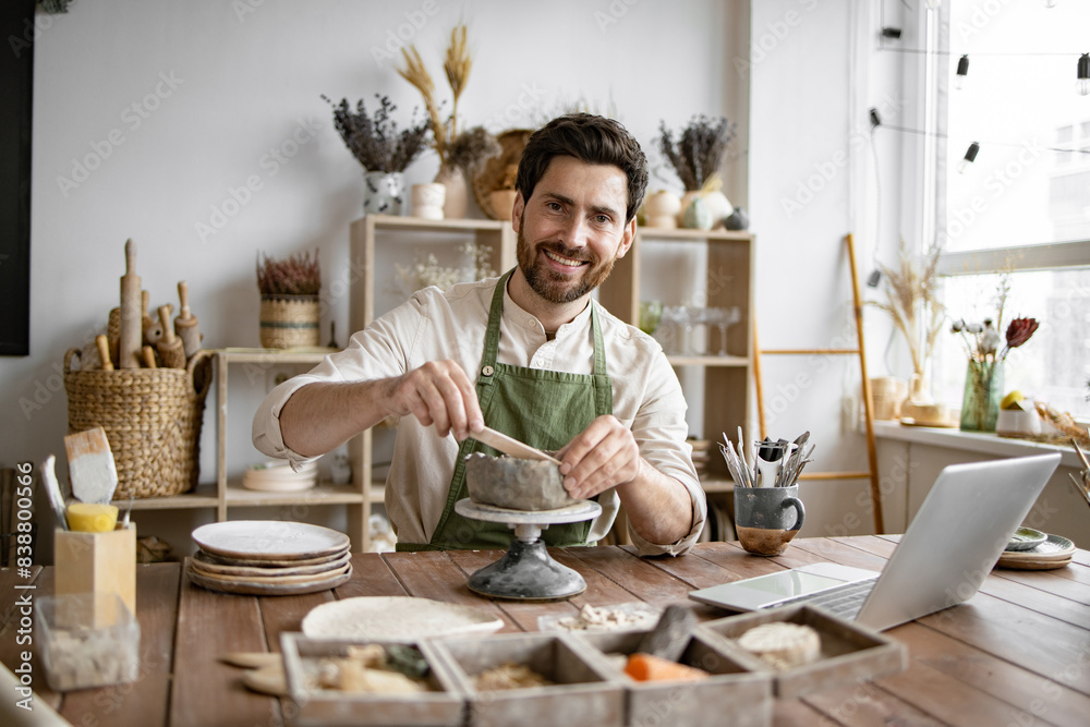 Bearded male seated at wooden table with various tools, materials, and laptop in bright well-decorated room. Smiling man working with clay in cozy home pottery studio. Bearded male seated at wooden table with various tools, materials, and laptop in bright well-decorated room. Smiling man working with clay in cozy home pottery studio.