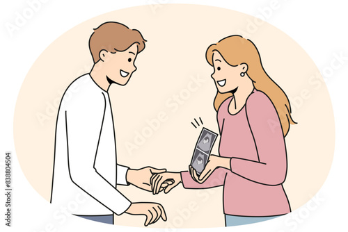 Pregnant woman holding ultrasound picture with baby showing future kiddy to happy father. Positive couple of man and girl getting ready to become parents and holding hands after going to doctor