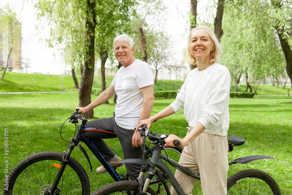 Fototapeta premium elderly senior couple rides bicycle in the park in the summer and smiles, old gray-haired man and woman are actively resting outdoors, old people practice cycling in forest