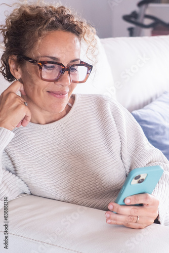 A happy curly lady with glasses relaxes at home alone, sits on the big sofa in a comfortable pose and shares good news on social media via mobile phone. Smiling woman enjoys weekend by ordering food 