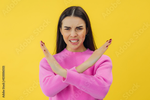 beautiful young woman unhappy holding two cross arms say no X sign, studio shot isolated yellow background, reject gesture