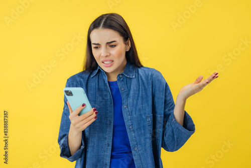 Annoyed angry young woman mad about spam message stuck phone looking at smartphone isolated on yellow studio background, furious teen girl having problem with cellphone irritated by broken mobile photo