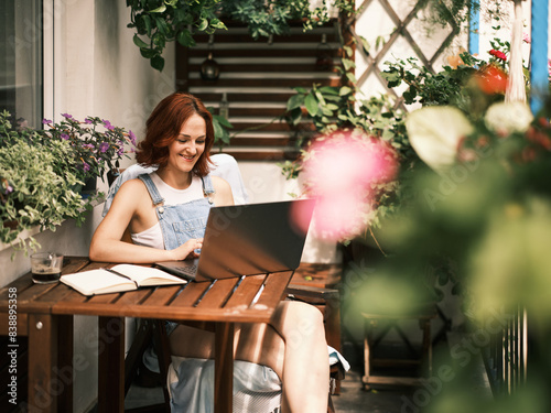 A smiling woman enjoys home gardening while using a laptop at an outdoor table surrounded by plants