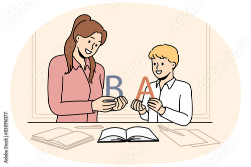 Back to school concept with boy learning alphabet along with elementary school teacher standing near table with books. Woman teaches child read or spell correctly, wanting give son quality education
