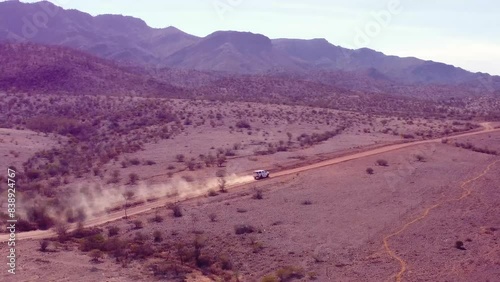 A white car kicks up dust as it speeds along a remote dirt road, leaving a trail of dust in its wake. The Rugged Gammon Ranges rise in the background, under a clear sky. photo