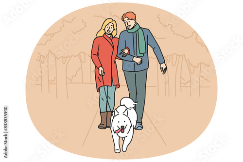 Man and woman are walking their dog together in park enjoying walk on autumn evening with warm weather. Happy couple with white dog on leash take care of pet in need of timely walk outdoors