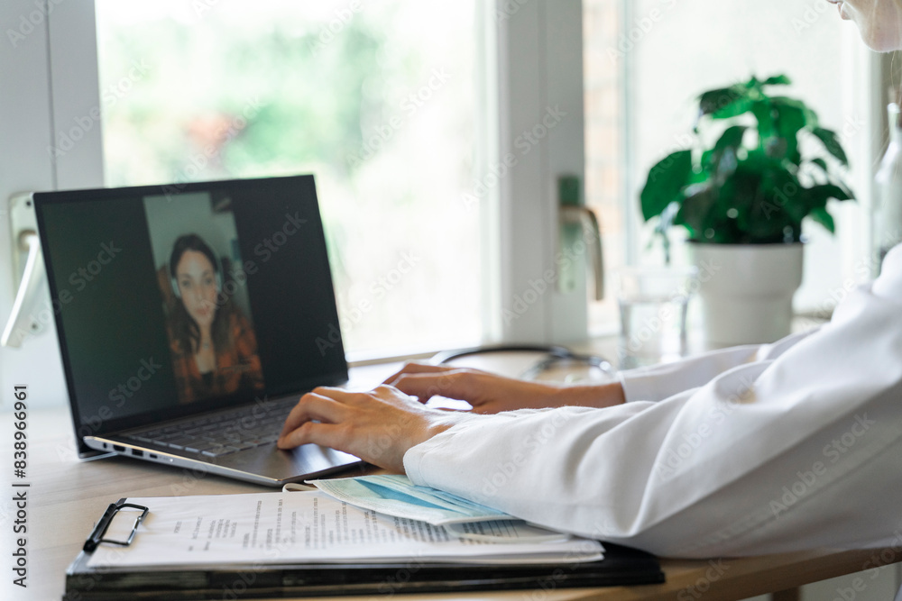 Fototapeta premium Young female doctor using laptop while talking to patient during online consultation
