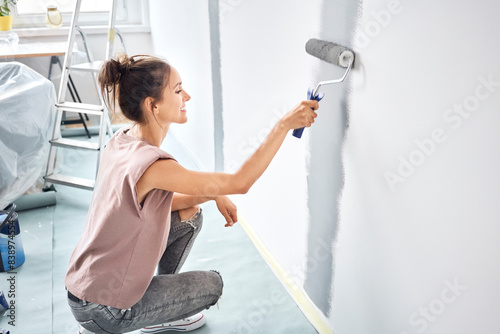Fototapeta Naklejka Na Ścianę i Meble -  Smiling woman painting with paint roller on wall while crouching at home