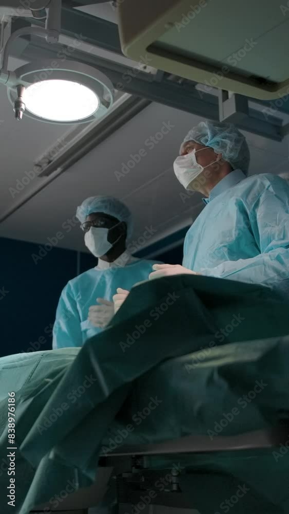 Two surgeons move an X-ray machine for surgery. Dark-skinned doctor in a mask and robe and his mature colleague prepare the patient for surgery. Vertical shot. Two surgeons move an X-ray machine for surgery. Dark-skinned doctor in a mask and robe and his mature colleague prepare the patient for surgery. Vertical shot.
