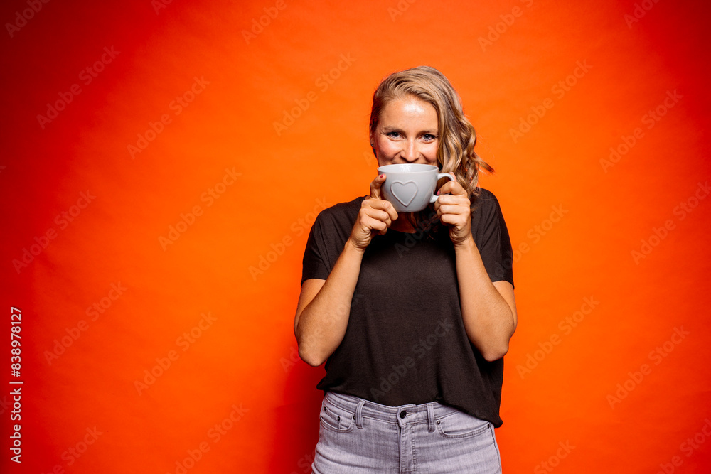 Happy woman holding coffee cup with heart shape while standing against orange background Happy woman holding coffee cup with heart shape while standing against orange background