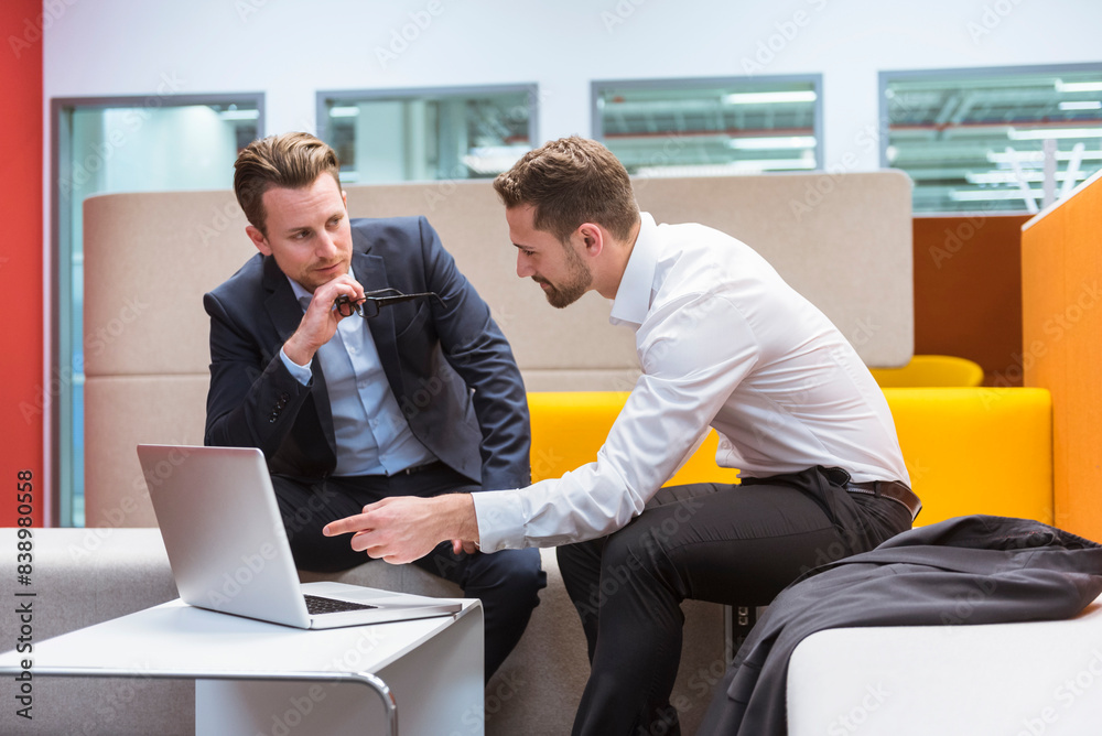 Businessmen sitting in conversation pit, discussing in front of laptop Businessmen sitting in conversation pit, discussing in front of laptop