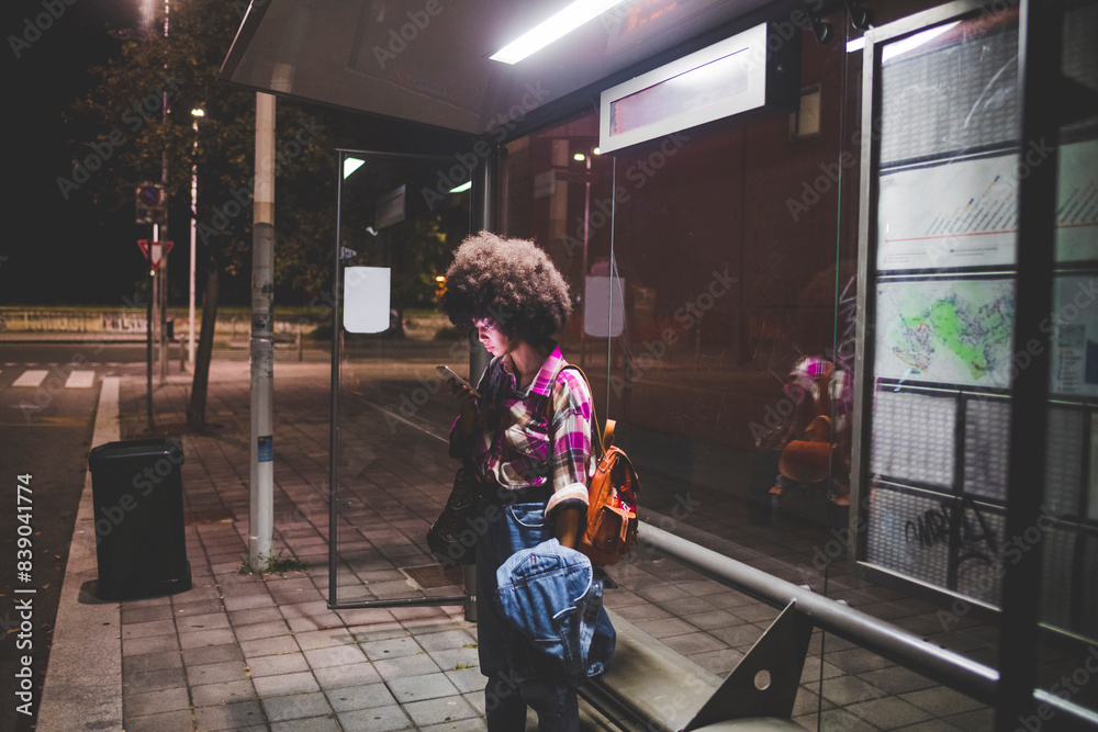 Young woman with afro hairdo using smartphone at bus stop in the city