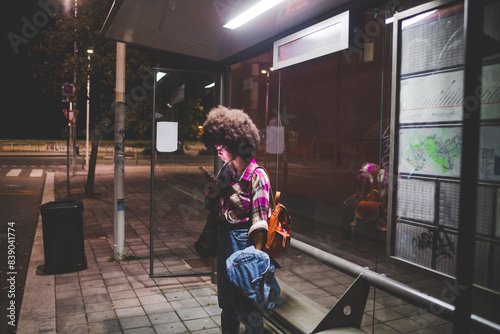 Young woman with afro hairdo using smartphone at bus stop in the city photo