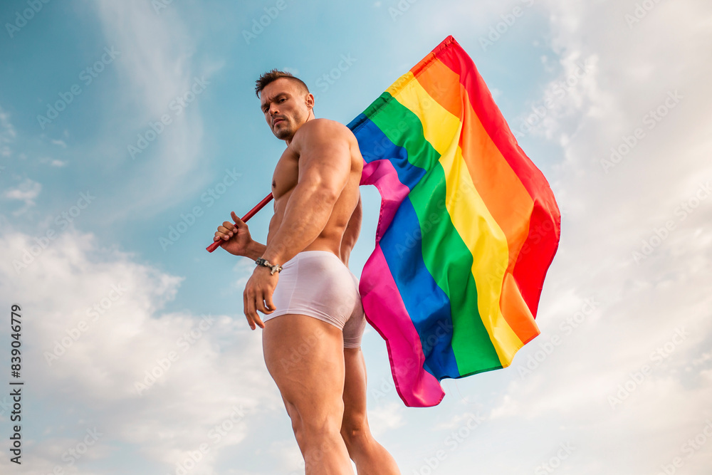 Shirtless young man holding rainbow flag while standing against sky
