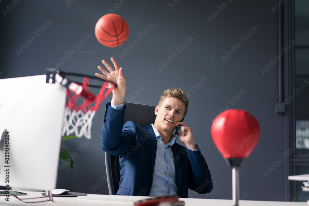 Young businessman sitting at desk in office playing basketball and talking on cell phone