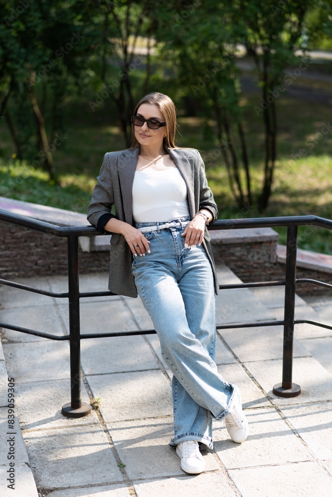 Portrait of a pretty and attractive Caucasian young woman in sunglasses and stylish casual clothes who is happy to pose while walking in the park on the steps on a warm spring day. Portrait of a pretty and attractive Caucasian young woman in sunglasses and stylish casual clothes who is happy to pose while walking in the park on the steps on a warm spring day.