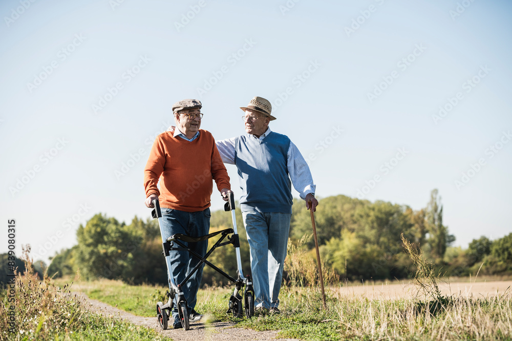 Fotografie Old friends taking a stroll in the fields with walking stick and wheeled walker,