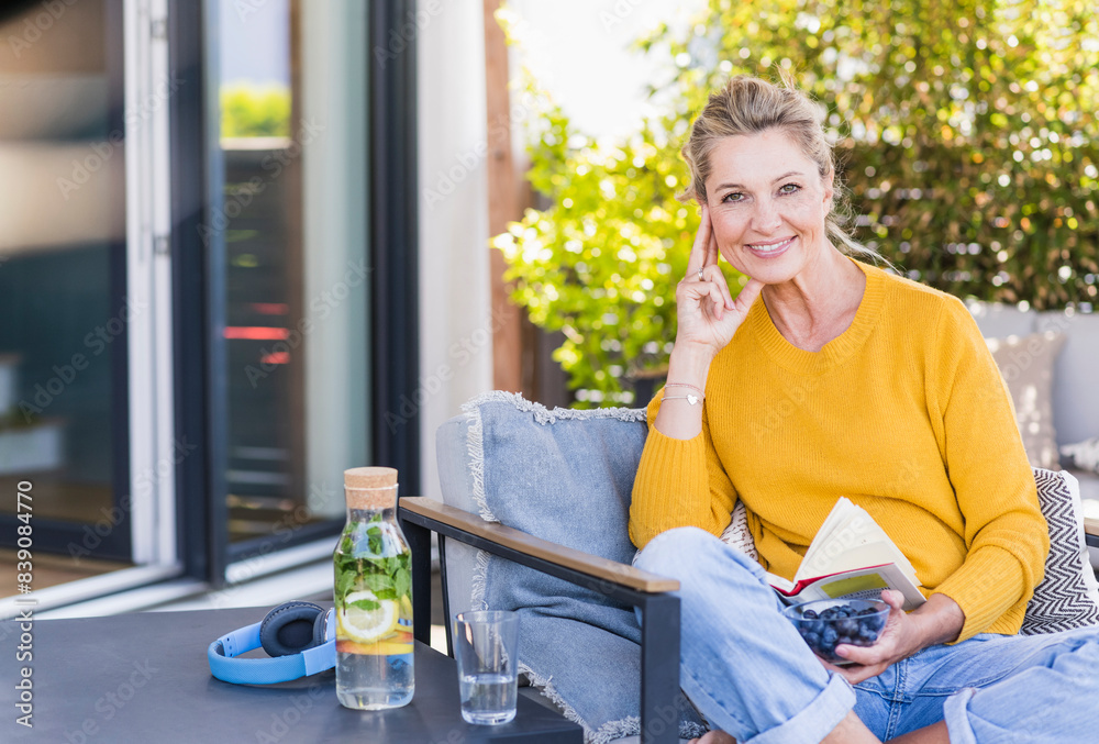 Portrait of smiling mature woman sitting on terrace with bowl of blueberries and book