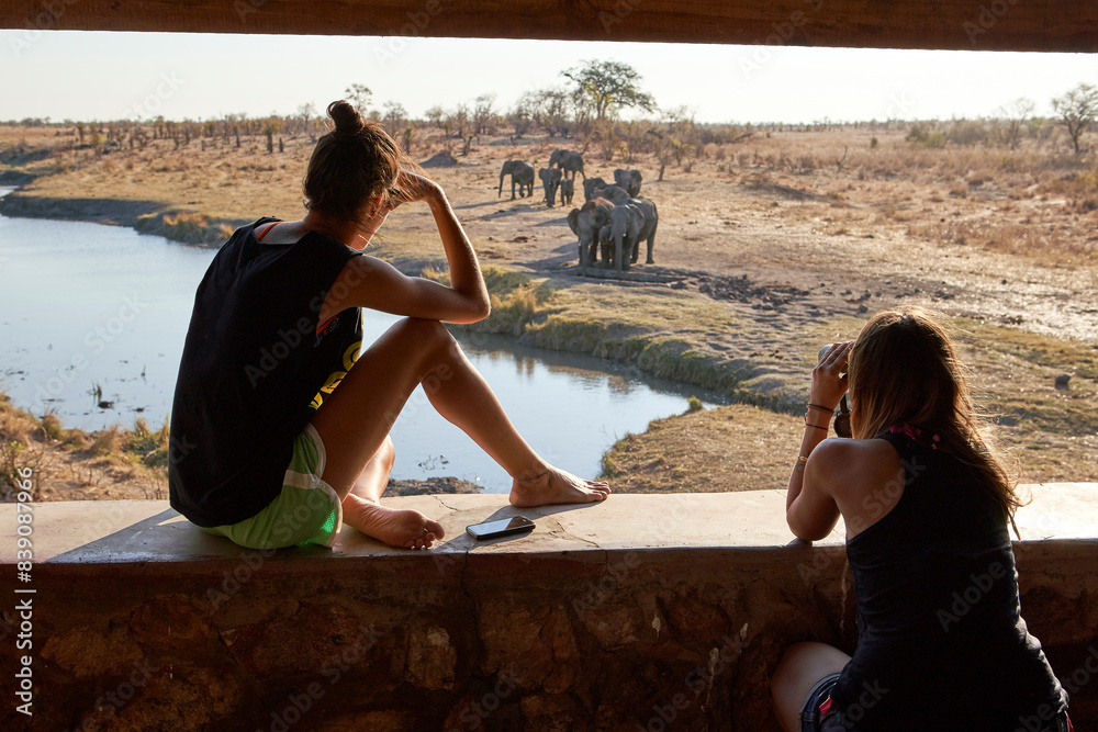 Two women watching a herd of elephants in the river from a viewpoint, Hwange National Park, Zimbabwe