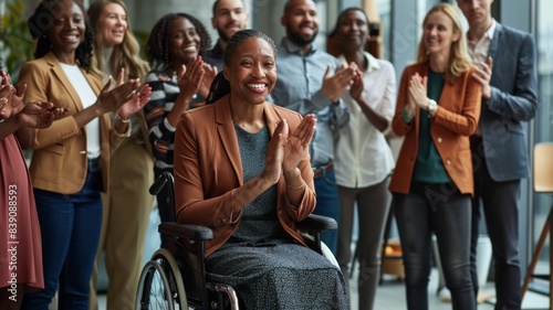 A group of business people are clapping and celebrating with one woman in a wheelchair. The modern office space shows a diverse group of employees standing around lady in the wheelchair cheering her