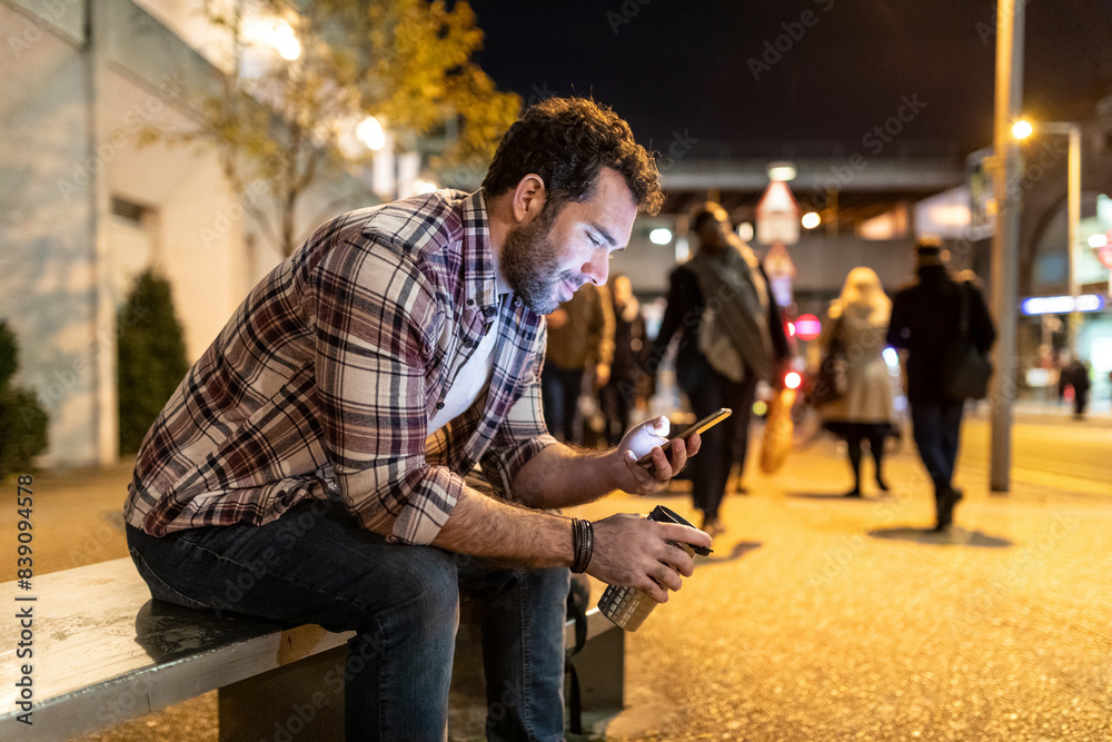 UK, London, smiling man sitting on a bench and looking at his phone by night