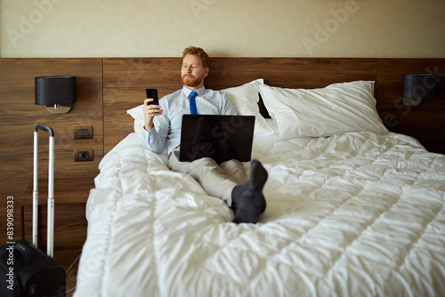 Obraz na płótnie Businessman lying on bed in hotel room using laptop and cell phone w salonie