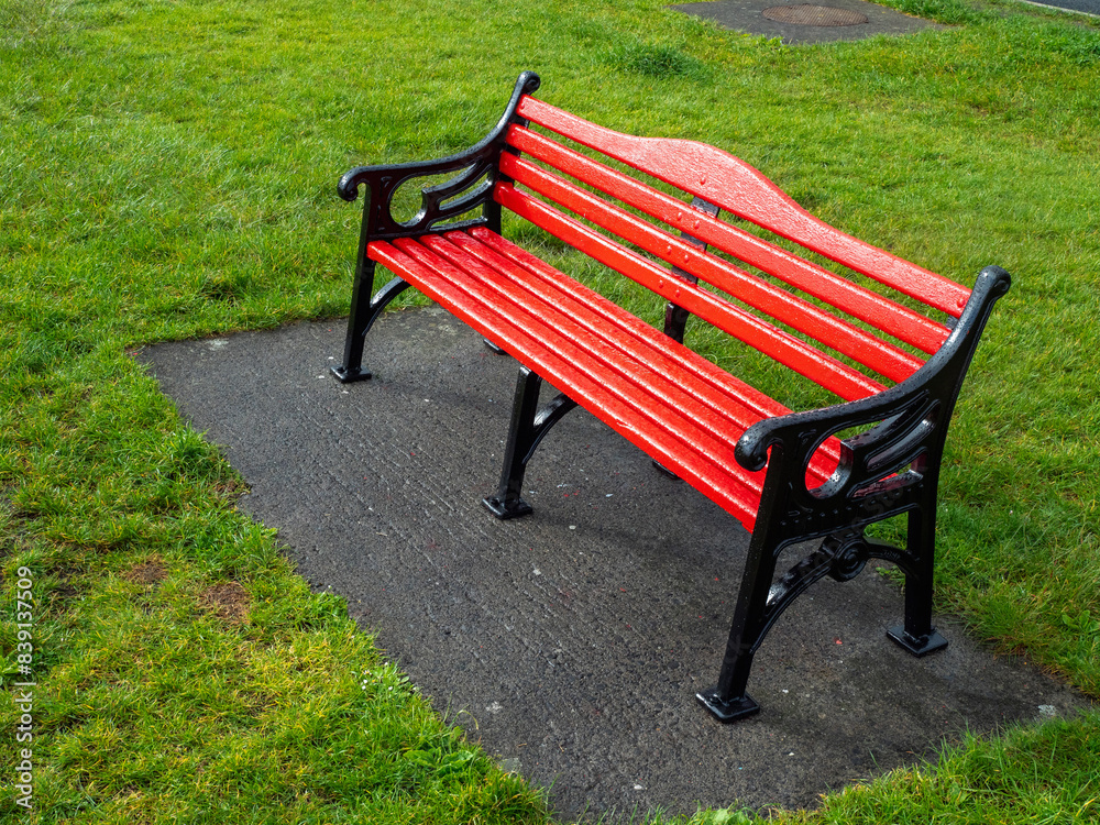 A red bench sits on a concrete slab in a grassy area. The bench is empty and the grass is lush and green. Classic metal cast old style design. Nobody. A red bench sits on a concrete slab in a grassy area. The bench is empty and the grass is lush and green. Classic metal cast old style design. Nobody.