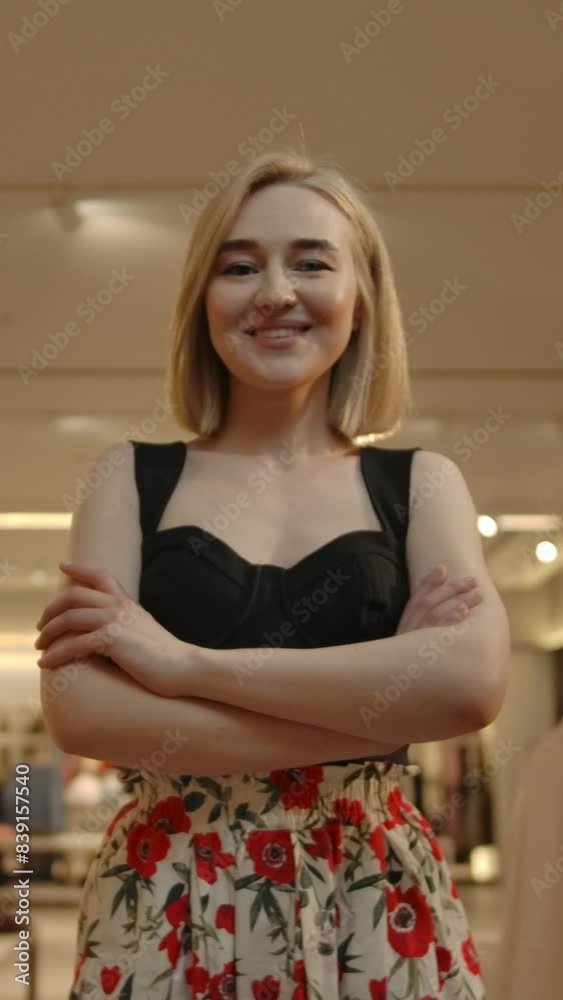 Zoom in view of happy young female crossing arms and smiling for camera while standing in modern store during weekend shopping. Vertical shot. Zoom in view of happy young female crossing arms and smiling for camera while standing in modern store during weekend shopping. Vertical shot.