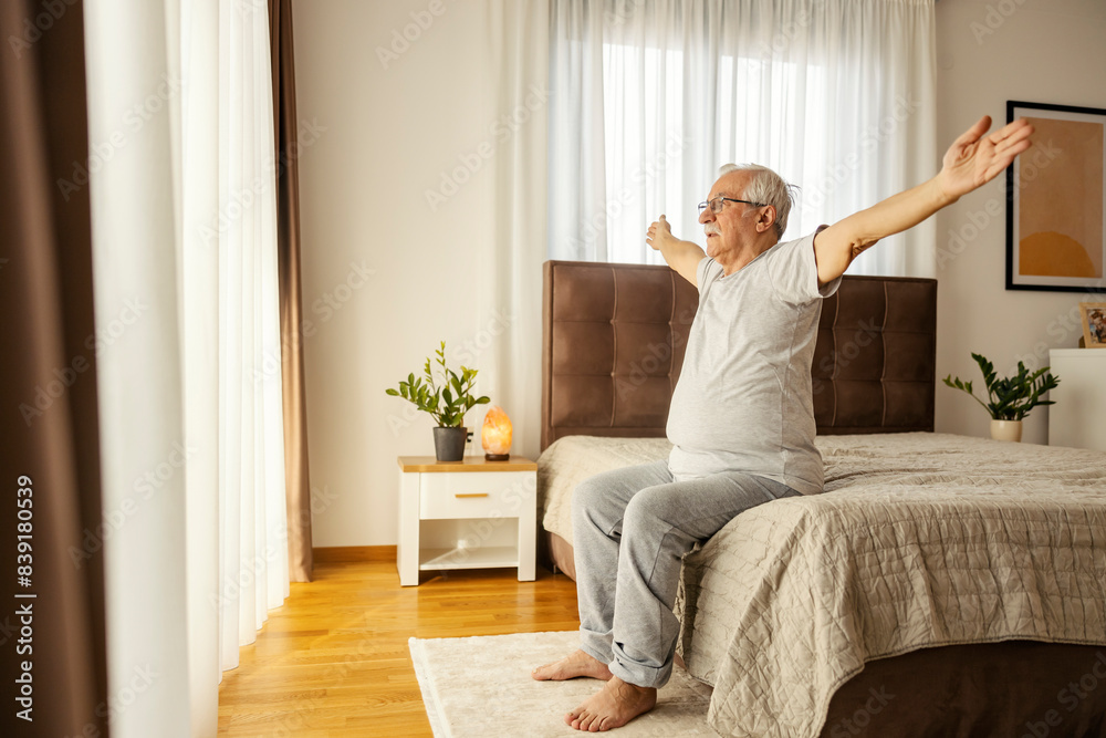 An active senior man in pajamas sitting on a bed in bedroom and doing morning workouts. An active senior man in pajamas sitting on a bed in bedroom and doing morning workouts.