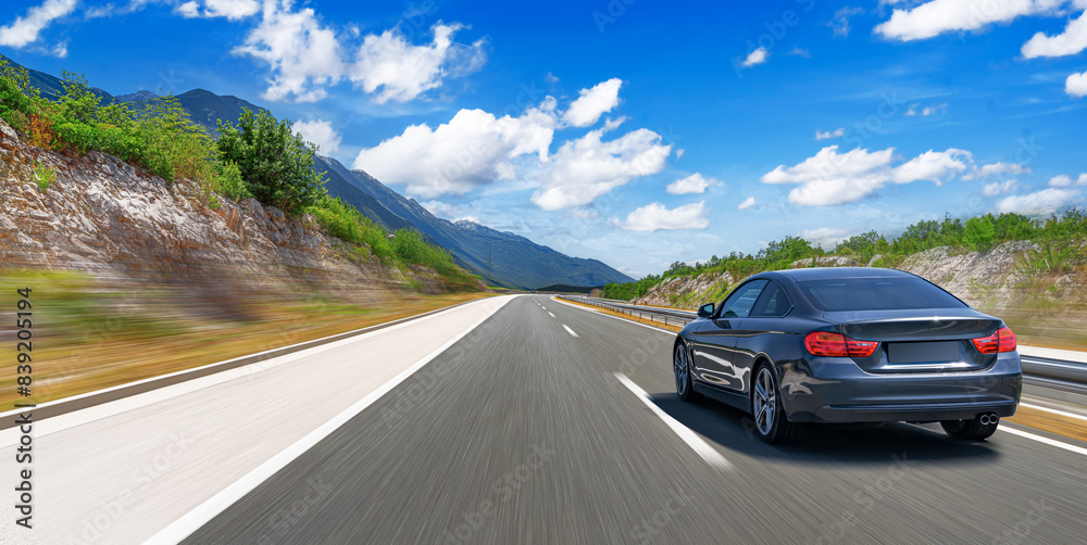 A grey car drives along the highway against the backdrop of rocky mountains on a sunny day.