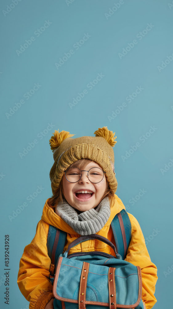 Young student boy in yellow jacket, glasses and a hat smiling with backpack in hand, isolated on blue background. Back to school concept. Copy space. Young student boy in yellow jacket, glasses and a hat smiling with backpack in hand, isolated on blue background. Back to school concept. Copy space.