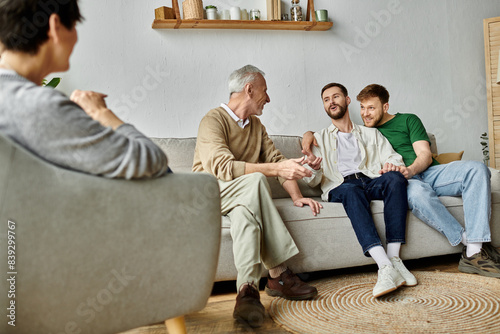A gay couple sits with parents on a couch, laughing and sharing stories.