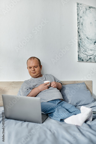 A man with inclusivity in pajamas relaxes in bed, holding a cup of coffee and looking at a laptop.