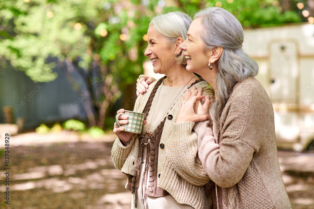 Fototapeta premium Two middle-aged women laugh together while enjoying a cup of coffee during a camping trip.